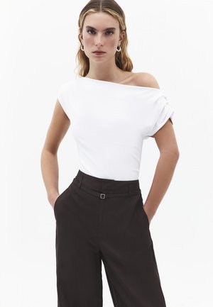 Kobieta o blond włosach, ubrana w biały top odsłaniający ramiona i czarne spodnie z wysokim stanem, stojąca z rękami w kieszeniach na białym tle.