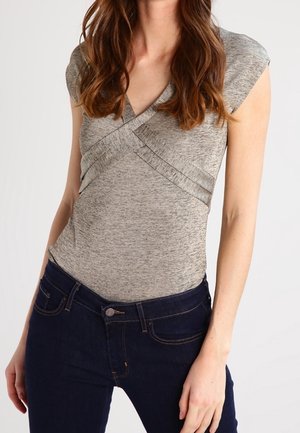 Vrouw draagt een grijs gewassen, korte mouw, overslagshirt en donkerblauwe spijkerbroek, staat tegen een eenvoudige lichte achtergrond.