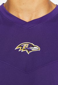 Fialový sportovní dres s výstřihem do V. Na hrudi se nachází logo Baltimore Ravens v žluté, černé a bílé barvě. Měkká, texturovaná tkanina.
