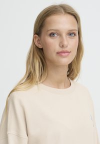 Sweat-shirt beige avec un col rond et des manches longues. Présente un petit logo bleu sur la poitrine. Tissu doux avec une texture lisse.