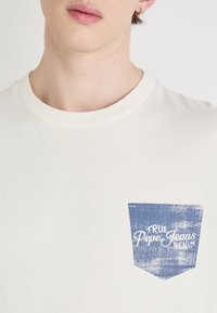 Kreminės spalvos marškinėliai su mėlynu spaudiniu kišenėje, ant kurio užrašyta "True Pepe Jeans Denim." Minkšta medžiaga, apvaliu kaklu.