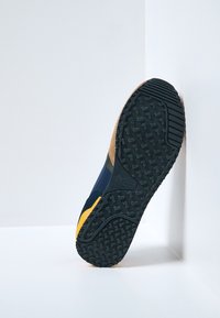 Semelle de chaussure en caoutchouc noir avec des motifs de grip, des accents jaunes et bleus, et une section beige. Conçue pour l'adhérence et la durabilité.