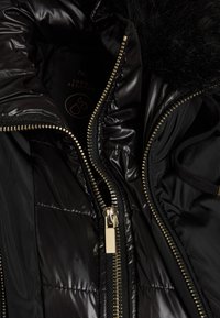 Czarna kurtka puchowa o błyszczącej, pikowanej strukturze, z detalami w złotym kolorze na zamku i kapturem wykończonym futrem. Posiada metkę "odporną na deszcz" w środku.