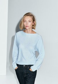 Giovane donna con capelli biondi corti, che indossa un maglione azzurro chiaro e jeans scuri, si trova con le mani in tasca su uno sfondo semplice.