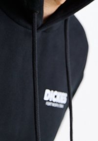 Hoodie noir en tissu doux, avec capuche à cordon de serrage et logo blanc "DICKIES" sur la poitrine. Le design est simple et fonctionnel.
