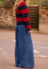 Suéter a rayas rojas y marino con cuello redondo, combinado con pantalones de mezclilla azul de pierna ancha. Enfoque en el corte relajado y la textura.