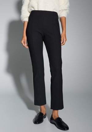 Frau trägt schwarze, figurbetonte, knöchellange Hose, schwarze Loafer und einen weißen, flauschigen Pullover, steht vor einem grauen Hintergrund.