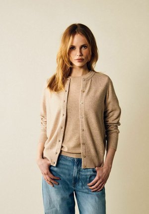 Beige cardigan med rund halsringning och knäppning, lagd över en matchande stickad topp, tillsammans med ljusblå jeans. Mjuk textur och avslappnad passform.