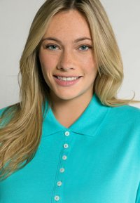 Tyrkysové polo tričko s límcem, obsahující šest perlizovaných knoflíků a texturovanou tkaninu. Pohled zblízka na límec.