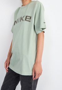 Mintgrön bomullströja med korta ärmar, försedd med en framträdande "NIKE"-logotyp i mörkgrått över framsidan. Klassisk avslappnad passform.