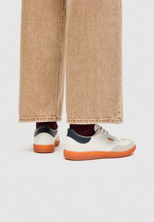 Persona, kas valkā bēša plata stila bikses, tumšas zeķes un baltas sporta kurpes ar tumšu papēža atzīmi un oranžu zolīti, stāv uz vienkārša fona.