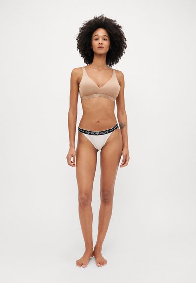 Bralette sans fil beige avec des fines bretelles et une bande texturée ; string crème assorti avec une ceinture logo noire.