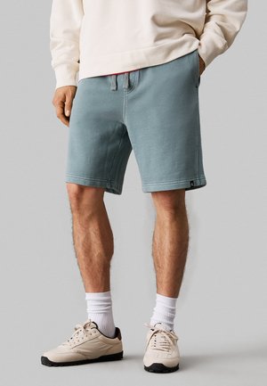 Pantalones cortos en tono azul desgastado, fabricados en un material suave, con una cinturilla elástica y cordón, bolsillos laterales y dobladillos cosidos. Zapatillas deportivas blancas completan el atuendo.