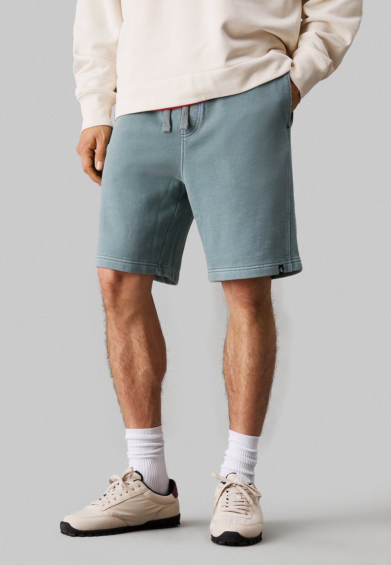 Pantaloni corti in teal sbiadito realizzati in tessuto morbido, con una fascia elastica in vita dotata di coulisse, tasche laterali e orli cuciti. Scarpe da ginnastica bianche completano l'outfit.