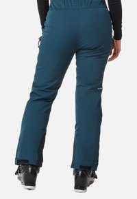 Türkisfarbene Skihosen mit einer glatten Textur, verstellbarem Bund und Reißverschlüssen an den Knöchellöchern, kombiniert mit schwarzen Skischuhen und auffälligen Beschlägen.