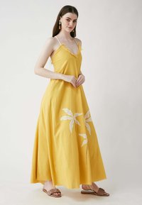 Gelbes Maxikleid mit Spaghetti-Trägern, Fransenbesatz und weißer Blattstickerei. Hergestellt aus leichtem Stoff, mit einer fließenden Silhouette.