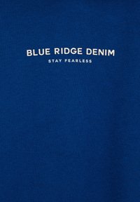 Kék szövet sima textúrával, amelyen fehér színű "BLUE RIDGE DENIM" és "STAY FEARLESS" felirat található, egyszerű, ívelt elrendezésben.