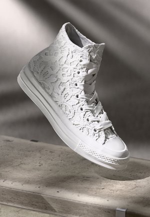 Witte hoge sneakers met kantbloemenpatroon en satijnen veters, zwevend boven een beige stenen oppervlak in zacht natuurlijk licht.