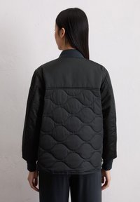 Schwarze gesteppte Jacke mit einem glatten, strukturierten Rücken. Sie hat gerippte Bündchen und einen Kragen, reicht bis zur Taille und verfügt über seitliche Reißverschlusstaschen.