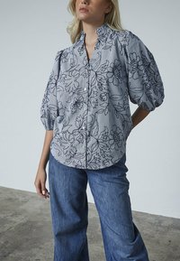 Stribet button-up skjorte med pufærmer, der har et blomsterprint i marineblå på en lyseblå baggrund, parret med brede denimjeans.
