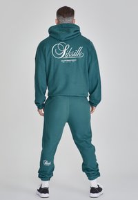 Mörkgrön hoodie och matchande joggers gjorda av mjukt tyg. Baksidan har vit märkestext. Ytterligare branding på joggersens ben.