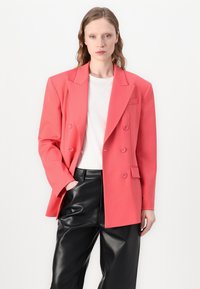 Koraalroze dubbelgeknopte blazer met getailleerde revers, voorzakken en een gladde textuur, gecombineerd met een witte T-shirt en zwarte neplederen broek.