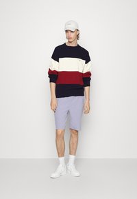 Maglione a righe orizzontali blu navy, rosso e bianco con un motivo testurizzato, abbinato a pantaloni corti viola chiaro e sneakers bianche. Cappellino bianco.