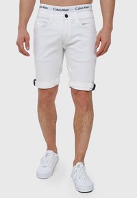 Shorts en denim blanc avec des ourlets retroussés, design à cinq poches, et une ceinture noire avec logo. Associés à des baskets blanches.
