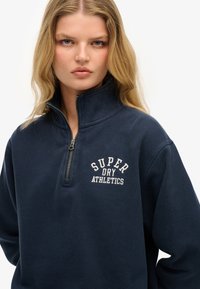 Sudadera azul marino con cuello de media cremallera, que presenta el texto "SUPER DRY ATHLETICS" en bordado blanco en el pecho. Tejido suave y texturizado.