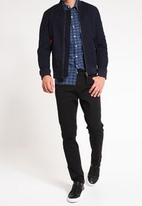 Blouson aviateur marine, chemise à carreaux bleue et noire, pantalon noir, et chaussures noires à enfiler avec semelles blanches. Design simple et coupe ajustée.