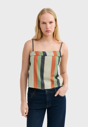 Femme aux longs cheveux blonds porte un haut sans manches à rayures verticales vertes, orange et bleu marine avec un jean bleu foncé, debout avec une main dans la poche.