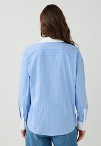 Vrouw in een lichtblauw gestreept shirt met een witte kraag en manchetten, gecombineerd met blauwe jeans, gezien vanuit de rug tegen een effen achtergrond.