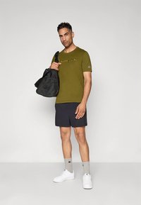 T-shirt justa verde-oliva com logótipo, calções pretos, ténis brancos, meias cinzentas e uma mala de ombro preta. Design simples, roupa casual.