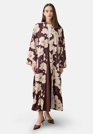 Kvinde iført en lang, mørk vinrød blomstret kimono-stil frakke over en hvid top og brun nederdel med spidse brune hæle.