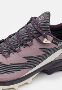 Trail-sko med en lila och grå stickad ovandel, texturerade överdrag, snörning och en framträdande Gore-Tex-tag på sidan.