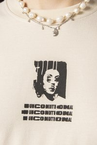 Koszulka w kolorze kremowym z czarnym nadrukiem graficznym przedstawiającym twarz oraz słowem „UNCONDITIONAL” powtarzanym na pogrubiony poniżej.