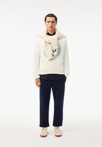Felpa crema con logo verde, pantaloni blu navy e un maglione beige drappeggiato attorno al collo. Scarpe color cammello con accenti chiari. Completo semplice e informale.