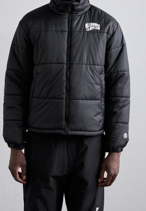 Homme portant une veste matelassée noire zippée avec le logo "Billionaire Boys Club" sur la poitrine et un pantalon noir, debout devant un fond uni.