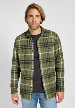 Vihreä ruudullinen flanellipaita mustilla ja valkoisilla raidoilla, nappikiinnitys, kaksi rintataskua ja pitkät hihat, käytetty mustan T-paidan päällä.