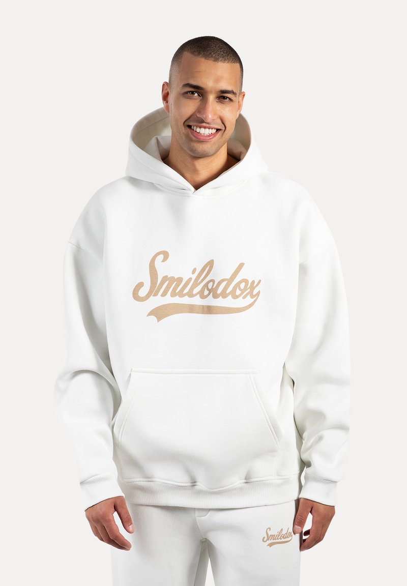 Sudadera blanca con un gran logo "Smilodox" en color beige. Presenta un bolsillo canguro, puños acanalados y una capucha forrada. Material suave y aterciopelado.