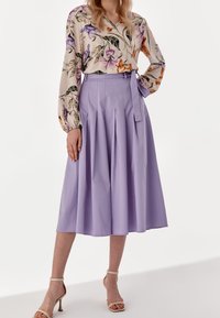 Kvinna klädd i en lila plisserad midikjol med knytbälte, krämfärgad blommönstrad blus med lång ärm och beige högklackade sandaler, stående mot vit bakgrund.