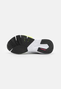 La suela en blanco y negro de una zapatilla deportiva presenta un patrón en zigzag, detalles en verde y la marca "PUMAGRIP" en rosa en la zona del talón.