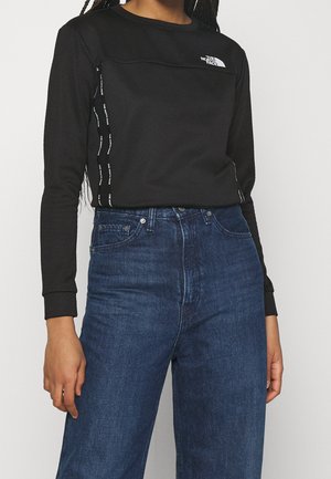 Person iført en sort North Face sweatshirt med lange ærmer og logo samt mørkeblå højhalsede jeans mod en ensfarvet baggrund.