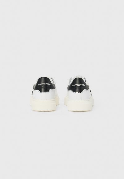 Białe skórzane sneakersy z czarnym, lakierowanym akcentem pięty i logo. Gruba, kremowa gumowa podeszwa, minimalistyczny design, błyszczące wykończenie.