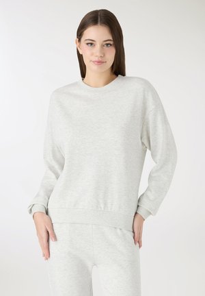 Giovane donna con capelli castani lisci che indossa una felpa grigio chiaro a maniche lunghe e pantaloni coordinati, in piedi davanti a uno sfondo bianco semplice.