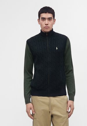 Jeune homme portant une veste noire en maille torsadée zippée sur une chemise à manches longues vert olive et un pantalon beige, debout devant un fond uni.