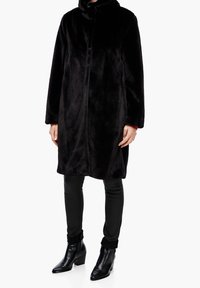 Cappotto in finta pelliccia nero con colletto alto, bottoni frontali e maniche lunghe; abbinato a pantaloni skinny neri e stivaletti alla caviglia. Texture liscia.