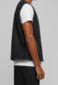 Czarna bezrękawnik z bawełny, o prostym, prostokątnym kroju z kieszenią z przodu i zaokrąglonym dołem, noszona na białej koszuli.