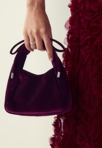 Geantă de mână din catifea mov cu o textură moale, cu două mânere legate și accente hardware argintii, ținută lângă o rochie texturată cu motive florale.