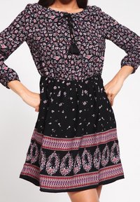 Vestido negro con flores, con un cuerpo fruncido, mangas largas y detalle de borlas. La falda presenta un dobladillo estampado en tonos de rosa y púrpura.